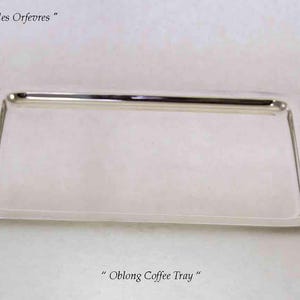 Könnte beinhalten: Ein poliertes, silberfarbenes, längliches Kaffeetablett mit glatter, reflektierender Oberfläche und abgerundeten Ecken. Der Text "Guilde des Orfevres" ist oben links sichtbar, und der Text "Oblong Coffee Tray" steht zentriert unter dem Tablett.