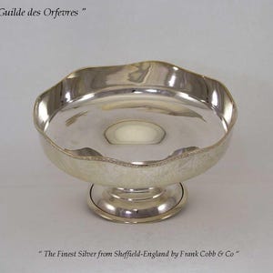 May include: A silver bowl with a wavy edge, standing on a short, round foot. The bowl has a wide, shallow basin. The text "Guilde des Orfevres" and "The Finest Silver from Sheffield-England by Frank Cobb & Co" are visible.