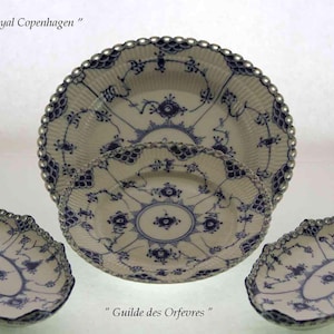 Puede incluir: Un conjunto de platos y fuentes de porcelana blanca con diseños florales azules y bordes plateados con cuentas. La imagen incluye dos platos grandes, un plato más pequeño y dos fuentes decorativas. El texto "The Pride of Royal Copenhagen" y "Guilde des Orfevres" son visibles.