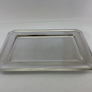 Könnte beinhalten: Ein rechteckiges silberfarbenes Serviertablett mit einer polierten, reflektierenden Oberfläche und einem breiten, flachen Rand mit abgeschrägten Kanten. Das Tablett hat eine saubere, glatte Oberfläche mit scharfen Ecken und einer vertieften Mitte.