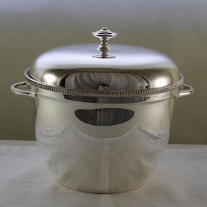 May include: A shiny silver ice bucket with a lid and two handles. The lid has a decorative finial. The bucket's surface reflects the surrounding environment. The rim of the bucket has a detailed design.