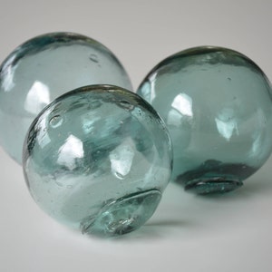 May include: Three glass fishing floats in a light blue-green color. The floats are round and smooth, with a slight sheen.