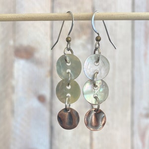 May include: Dangling earrings with a unique design. Each earring features three iridescent, round, button-like elements connected by small metal rings. The bottom element is a small, hammered copper disc. The earrings hang from silver-colored ear wires.