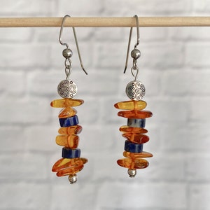 May include: A pair of dangle earrings with silver hooks and a silver charm at the top. The earrings feature a stack of amber and blue lapis lazuli beads.