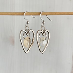 May include: A pair of silver heart-shaped earrings with a hammered texture. Each earring features a small, oval-shaped, off-white stone suspended within the heart. The earrings hang from silver ear wires.