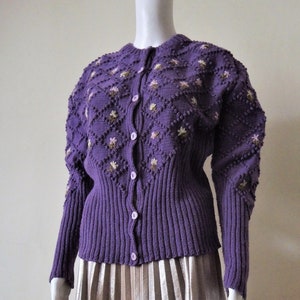 May include: A purple knitted cardigan with a floral embroidered design and button closure. The cardigan has a fitted silhouette and long sleeves.