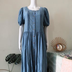 Könnte beinhalten: Ein hellblaues Jeans-Midikleid mit kurzen Puffärmeln, einem Rundhalsausschnitt und einer durchgehenden Knopfleiste auf der Vorderseite. Das Oberteil ist mit vertikalen Biesen auf beiden Seiten der Knopfleiste versehen.