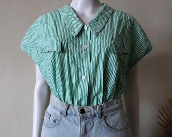 May include: A green and white striped short-sleeved button-up shirt with a collared neckline and two chest pockets. The shirt is worn with light blue denim shorts.