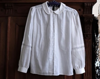 May include: White collared blouse with long sleeves and lace trim. The blouse is hanging on a wooden hanger against a dark wood background.