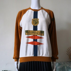 Puede incluir: Una sudadera de manga larga blanca y marrón con un estampado gráfico de una cabeza de tigre y el texto "Chitawan National Park".