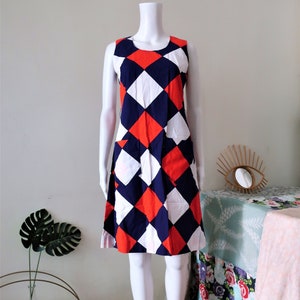May include: A sleeveless dress with a geometric pattern in red, white, and blue. The dress has a diamond pattern and two pockets.