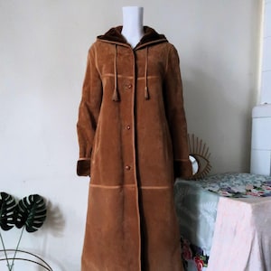 May include: A long brown suede coat with a hood and a drawstring. The coat has a button closure and a tassel detail on the drawstring.