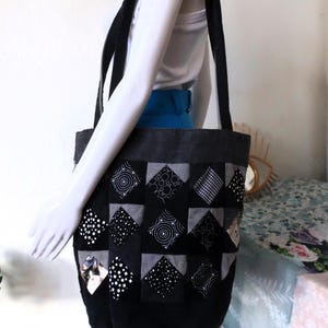 Può includere: Una borsa tote nera in patchwork con una varietà di motivi in ​​bianco e nero. La borsa ha due manici neri ed è realizzata in un tessuto resistente.