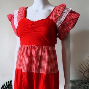 Op de afbeelding: Rood en witte patchwork jurk op een mannequin. De jurk heeft een vierkante halslijn en gefranjerde mouwen. Het lijfje is effen rood, het middenstuk heeft een ruitpatroon en de rok heeft stippen.