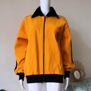 May include: A yellow zip-up jacket with black trim and a black collar. The jacket has a small embroidered patch on the chest with a red and yellow design.