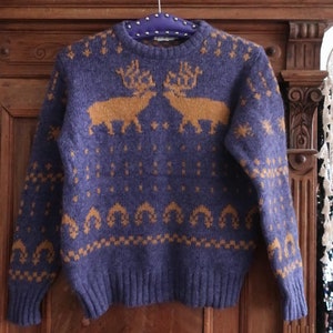 Puede incluir: Un suéter de punto azul con un patrón de reno marrón. El suéter tiene cuello redondo y mangas largas.