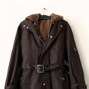 Op de afbeelding: Een donkerbruine jas met een capuchon en een riem. De capuchon heeft een bruine, fuzzy voering. De jas heeft een knoopsluiting en zakken. De jas hangt aan een zwarte hanger tegen een witte achtergrond.