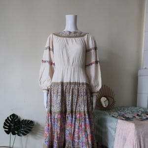 May include: A long, white, tiered dress with floral print and a gathered bodice. The dress has long sleeves with floral trim and a round neckline.