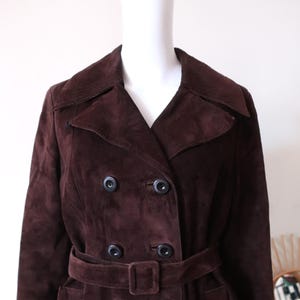 May include: A long brown suede coat with a double breasted closure, a belt, and pockets.