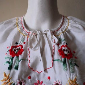 Könnte beinhalten: Weiße Baumwollbluse mit bunten gestickten Blumen, darunter rote Mohnblumen, blaue Kornblumen und gelber Weizen. Die Bluse hat einen gerafften Ausschnitt mit einer weißen Schleife.