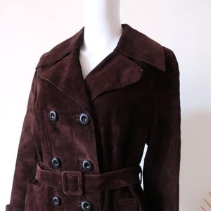 May include: A long brown suede trench coat with a double breasted closure, a belt, and a collar.