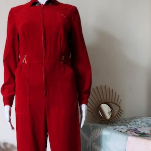 May include: A red corduroy jumpsuit with a zipper closure and long sleeves. The jumpsuit has a collared neckline and two pockets on the front.
