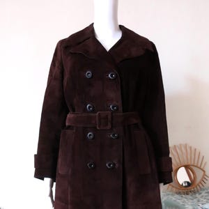 May include: A long brown suede coat with a double breasted closure, a belt, and two pockets.