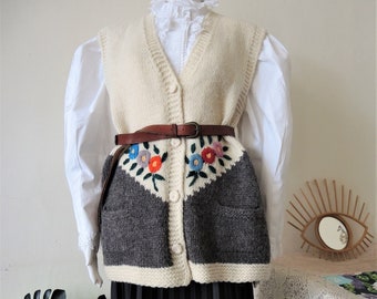 Könnte beinhalten: Eine cremefarbene Strickweste mit einem braunen Ledergürtel und bunter Blumenstickerei. Die Weste hat einen V-Ausschnitt und einen Knopfverschluss. Der untere Teil der Weste ist grau und weiß.