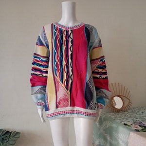 May include: A colorful sweater with a patchwork design. The sweater features a mix of patterns and colors, including pink, blue, yellow, and white. The sweater has long sleeves and a crew neck. The garment is displayed on a mannequin.