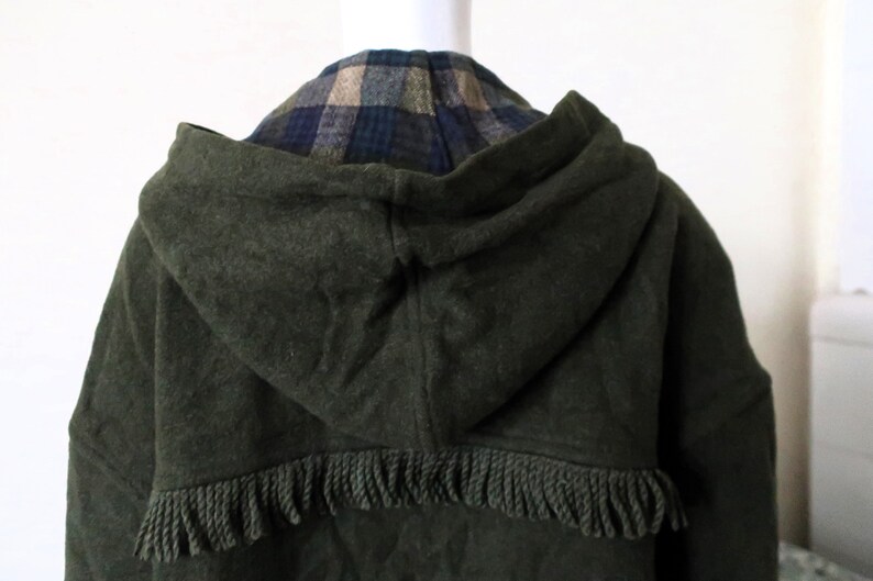 May include: Dark green hooded coat with a fringed hem. The hood reveals a plaid pattern in shades of blue, tan, and green. The coat is made of a thick, textured fabric.