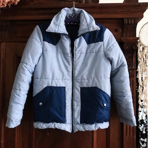May include: A vintage blue and white checked jacket with navy blue panels and pockets. The jacket has a full zip, a classic collar, and is displayed against a dark wooden background. The jacket is a retro style.