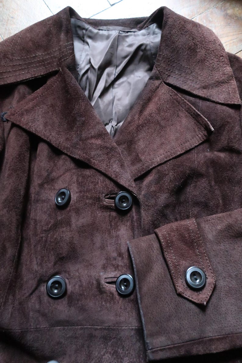 May include: A brown suede jacket with a double breasted closure and a large collar. The jacket has four black buttons.