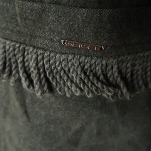 May include: Close-up of a dark green wool garment with a decorative fringe along the edge. The garment features a small metal tag with the brand name "GEIGER". The fringe is a darker shade of green.
