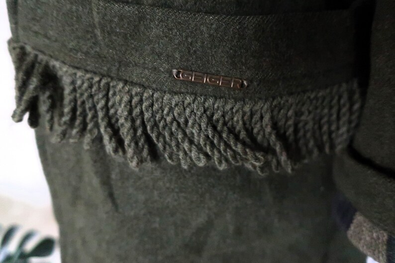 May include: Close-up of a dark green wool garment with a decorative fringe along the edge. The garment features a small metal tag with the brand name "GEIGER". The fringe is a darker shade of green.