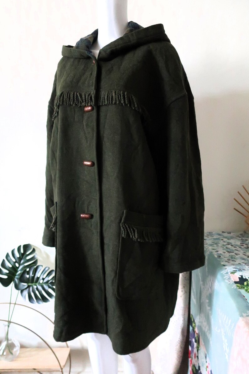 May include: Dark green hooded coat with fringe detailing and wooden toggle closures. The coat features two front pockets with fringe and a plaid lining in the hood. A vintage style coat.