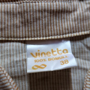 Może przedstawiać: Biała metka z tekstem "vinetta 100% BOMULL 38" w ż&oacute;łtych literach. Metka jest przymocowana do brązowo-białej tkaniny w paski.