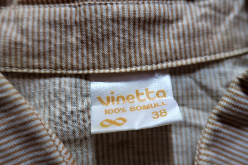 Może przedstawiać: Biała metka z tekstem "vinetta 100% BOMULL 38" w ż&oacute;łtych literach. Metka jest przymocowana do brązowo-białej tkaniny w paski.