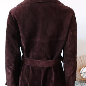 May include: A long, brown suede coat with a belt. The coat has a classic trench coat style with a collar and a double-breasted closure.