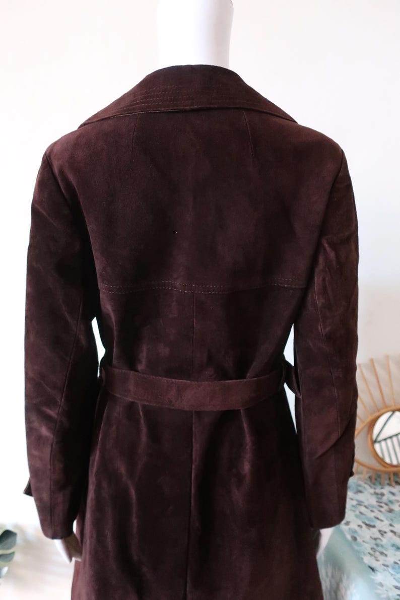 May include: A long, brown suede coat with a belt. The coat has a classic trench coat style with a collar and a double-breasted closure.