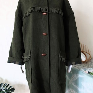 May include: Dark olive green hooded coat with fringe detailing across the chest and on the pockets. The coat has wooden buttons and a plaid lining on the cuffs and hood. A vintage style coat.