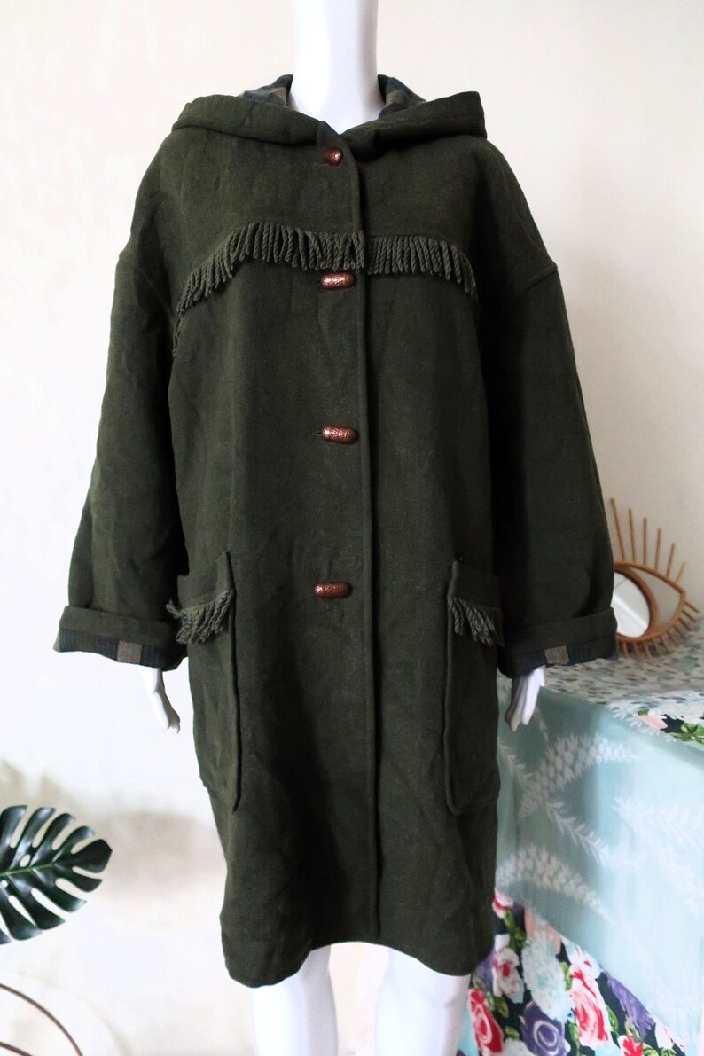 May include: Dark olive green hooded coat with fringe detailing across the chest and on the pockets. The coat has wooden buttons and a plaid lining on the cuffs and hood. A vintage style coat.
