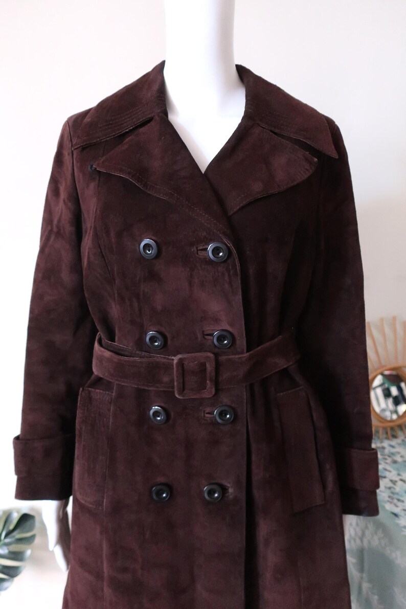 May include: A long brown suede coat with a double breasted closure, a belt, and pockets.