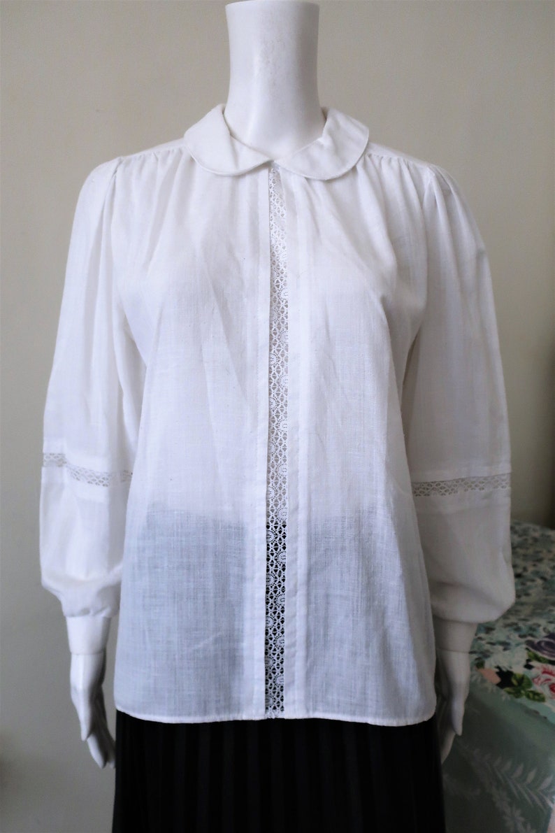 May include: A white cotton blouse with a peter pan collar and lace trim down the front and on the sleeves.