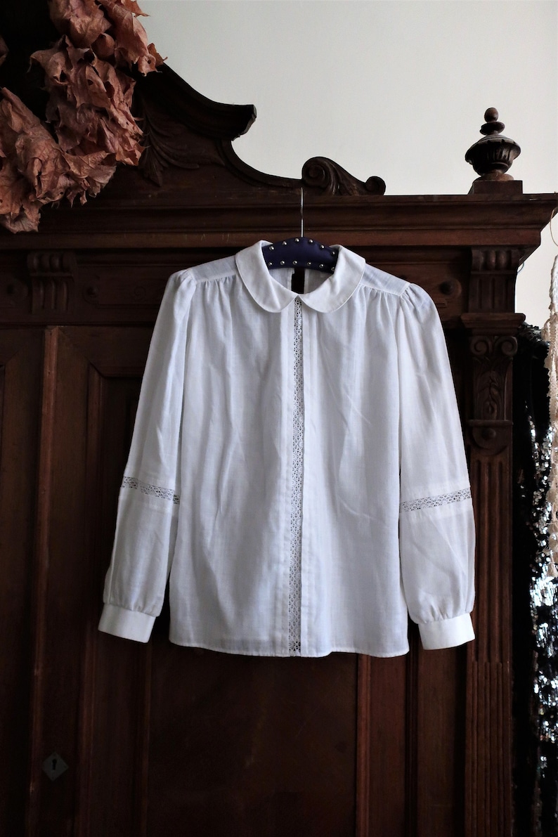May include: White collared blouse with long sleeves and lace trim. The blouse is hanging on a wooden hanger against a dark wood background.