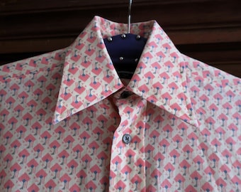May include: A button-down shirt with a repeating pattern of pink and blue shapes on a cream background. The shirt has a wide collar and a single button closure. The shirt is hanging on a hanger.