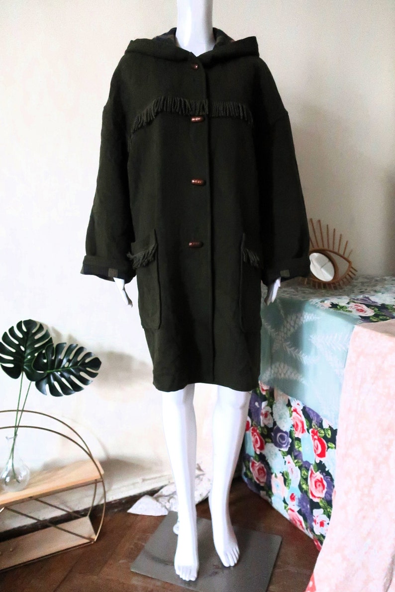May include: Dark olive green hooded coat with fringe detail on the chest and pockets. The coat has three brown buttons and a relaxed fit. The sleeves are cuffed, and the coat is displayed on a mannequin.