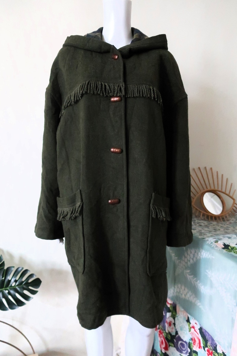 May include: Dark green hooded coat with fringe detailing and wooden buttons. The coat features two front pockets with fringe and a hood. The coat is a knee-length style, suitable for cooler weather.