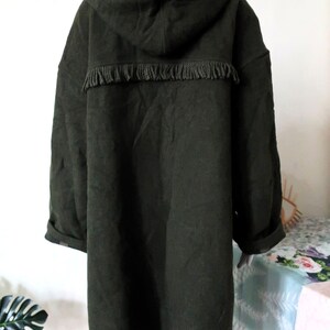 May include: Dark green hooded coat with fringe detail across the upper back. The coat has long sleeves and a relaxed fit. The hood and the inside of the coat's collar feature a plaid pattern. The coat is displayed on a mannequin.