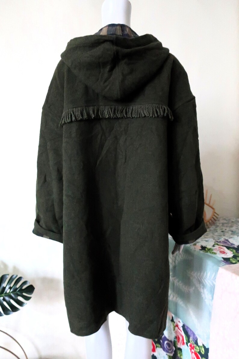 May include: Dark green hooded coat with fringe detail across the upper back. The coat has long sleeves and a relaxed fit. The hood and the inside of the coat's collar feature a plaid pattern. The coat is displayed on a mannequin.