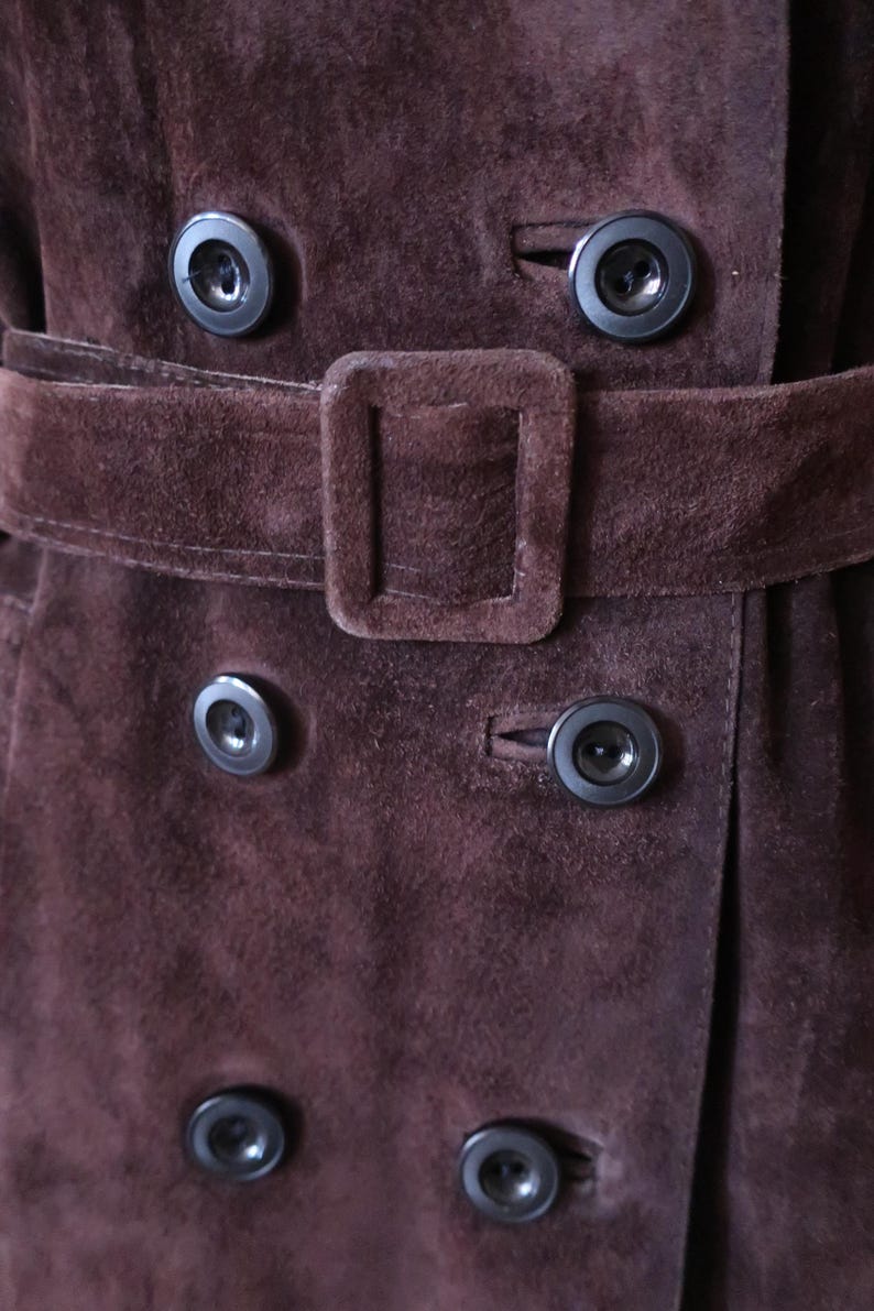 May include: Close-up of a brown suede jacket with a belt and multiple black buttons.